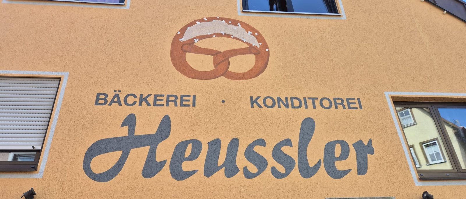 Hausfassade der Bäckerei Heussler: eine große gemalte Brezel über dem Schriftzug „Bäckerei · Konditorei Heussler“.
