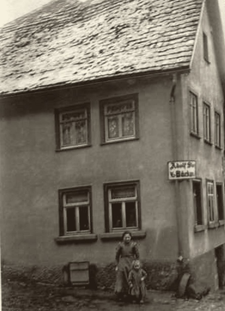 Altes Bäckerhaus mit schwerem Ziegeldach; eine Frau mit Kind steht vor der Hauswand.